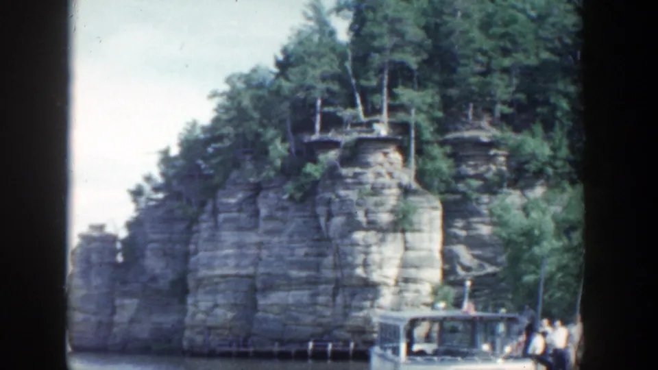 1964 Wisconsin 8mm frame showing a river tour boat near a tree-covered limestone cliff under a pale sky.