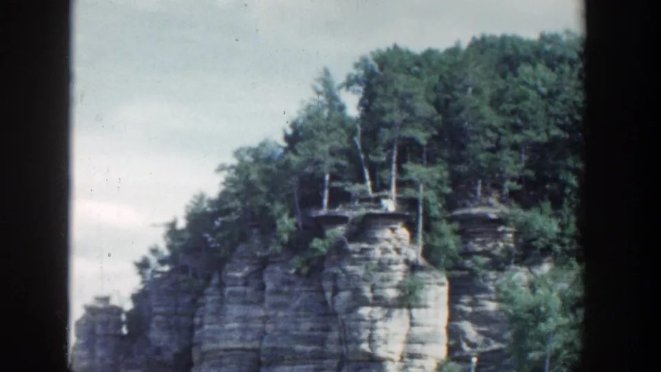 1964 Wisconsin home-movie frame of a limestone bluff with trees above the river, filmed from a boat perspective.