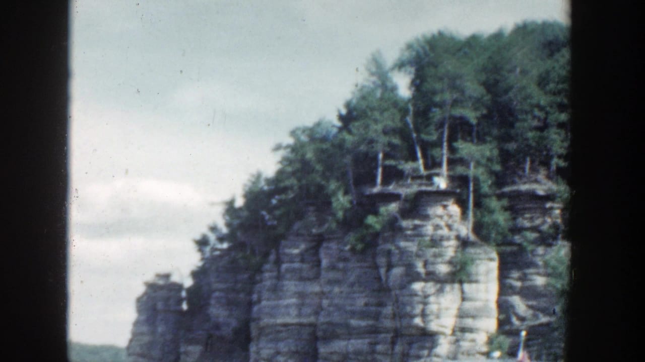 1964 Wisconsin 8mm view of tree-topped layered rock rising from calm water, framed by dark film edges.