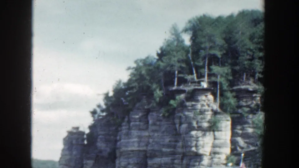 1964 Wisconsin 8mm view of tree-topped layered rock rising from calm water, framed by dark film edges.