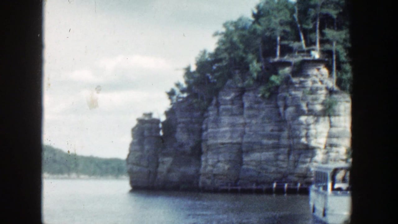 Rocky cliff edge and wooded shoreline beside open water, with part of a boat at right.