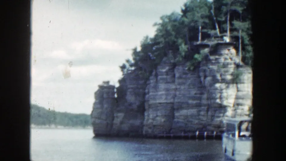 1964 Wisconsin 8mm frame showing a cliff face, trees, open water, distant shore, and a partial boat edge.