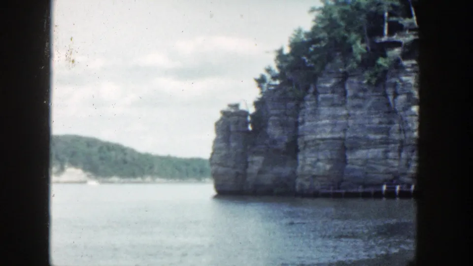 1964 Wisconsin 8mm view of a stratified rock wall beside calm water, with trees and distant shore visible.