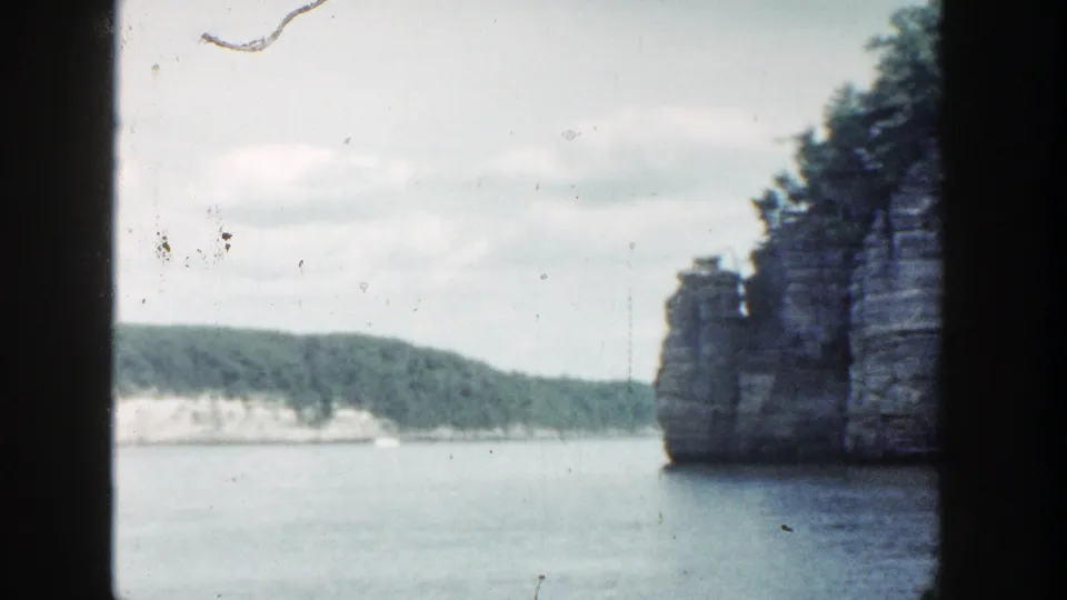 1964 Wisconsin 8mm frame with open water, distant tree-lined shore, pale sky, and a rock wall at right.