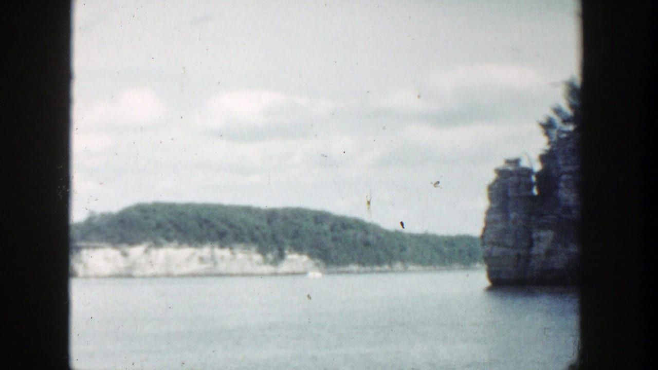 1964 Wisconsin 8mm frame showing calm water, distant rocky shoreline, trees, cloudy sky, and heavy gate-edge borders.