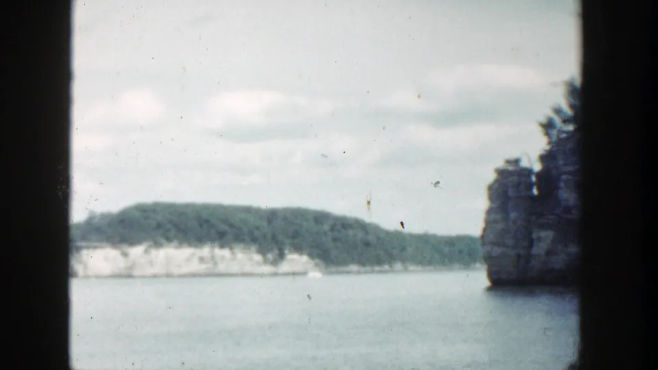 1964 Wisconsin 8mm frame showing calm water, distant rocky shoreline, trees, cloudy sky, and heavy gate-edge borders.