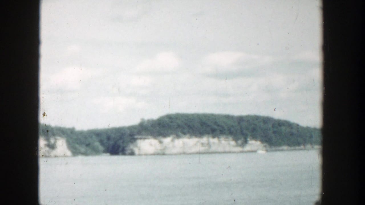 1964 Wisconsin lake view from a boat, with a dark vertical film border dividing rocky wooded shoreline and distant pale cliffs.
