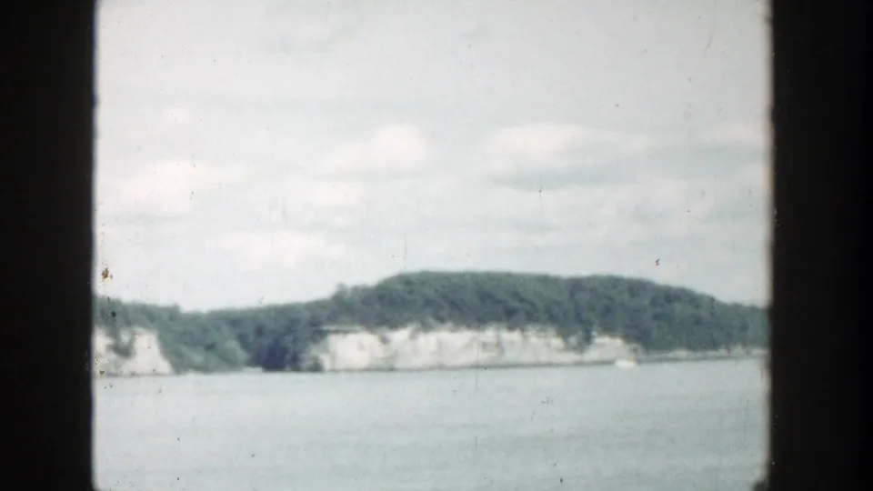 1964 Wisconsin lake view from a boat, with a dark vertical film border dividing rocky wooded shoreline and distant pale cliffs.