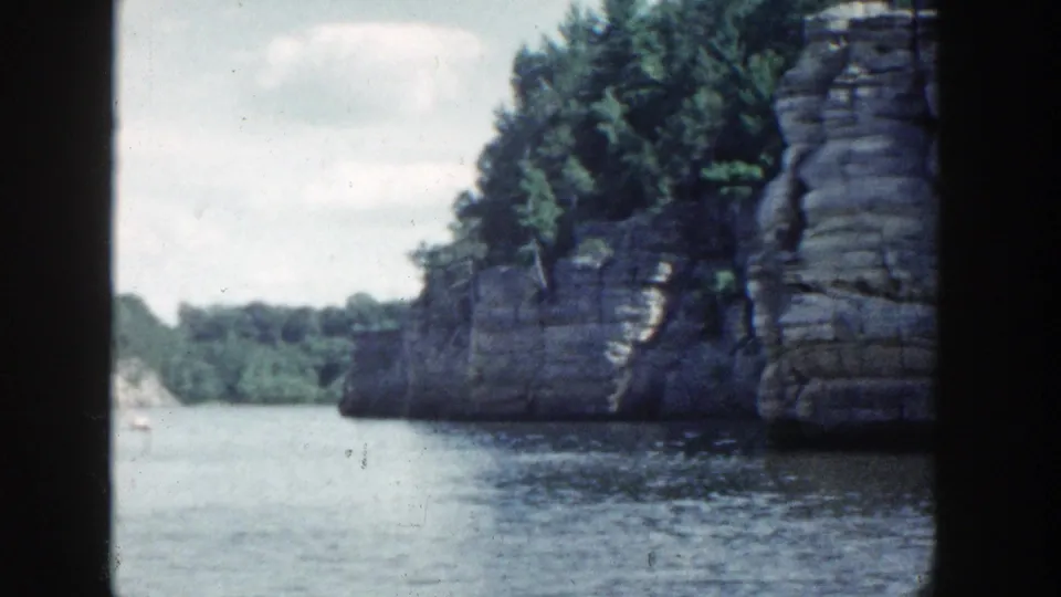 Close 1964 Wisconsin 8mm view of layered shoreline rocks, evergreen trees, lake surface, and visible film wear.