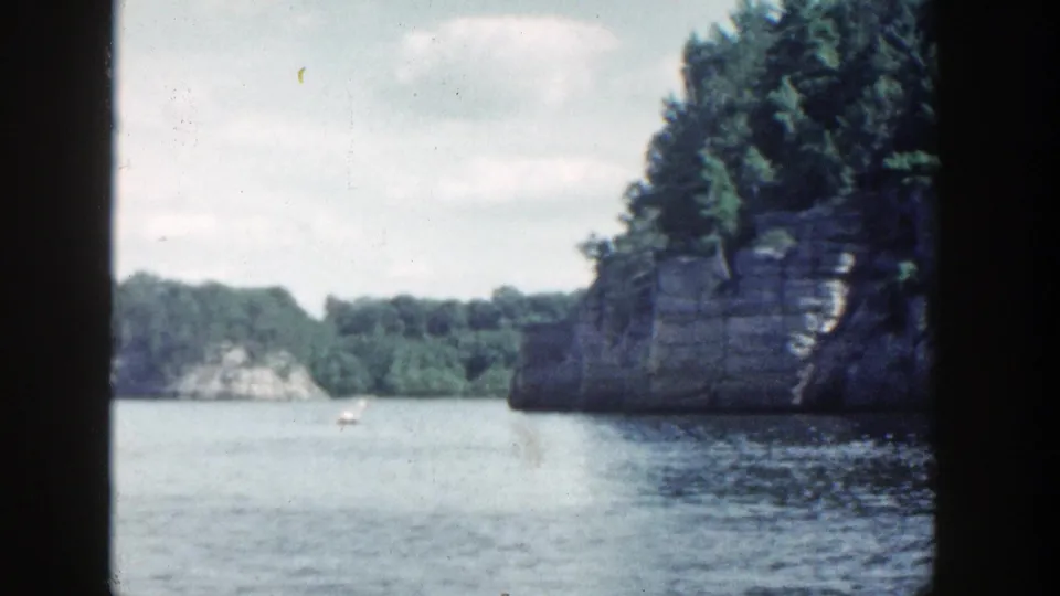 1964 Wisconsin home-movie frame showing a rocky wooded outcrop by the lake, distant shore, cloudy sky, and 8mm film grain.