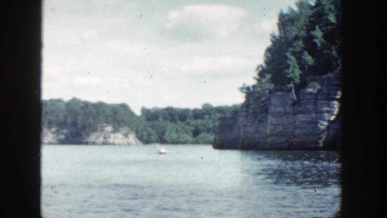Lake view with rocky wooded cliffs, calm water, dark frame edges, and a small distant boat.