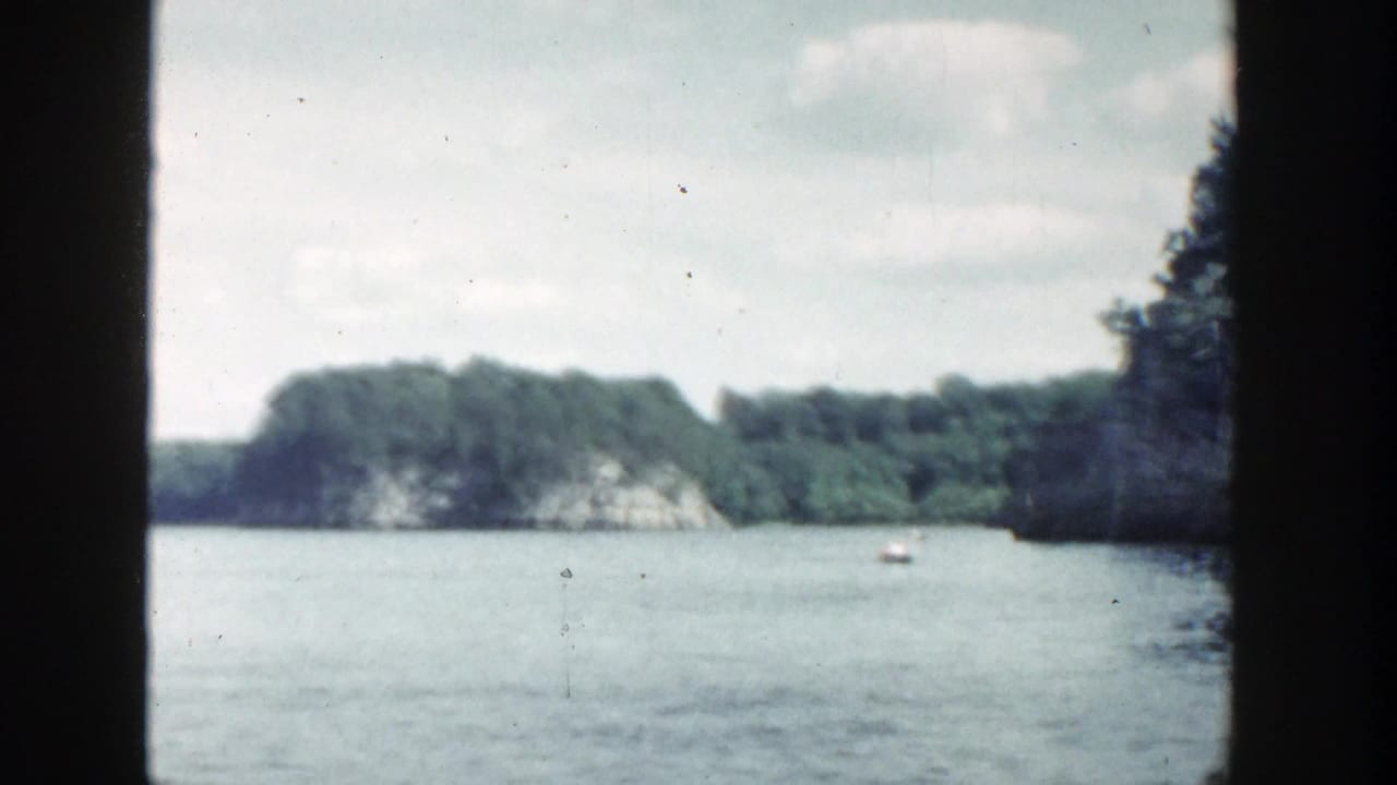 Wide lake scene with wooded cliffs, pale sky, small boat, and dark vertical film-edge framing.
