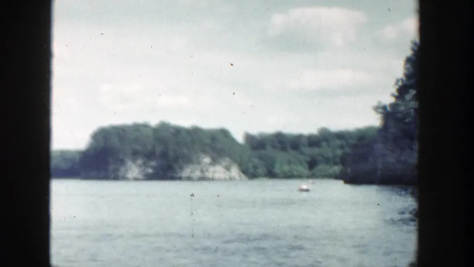 1964 Wisconsin lake view with a small white boat near wooded rocky shoreline, seen through dark boat or window framing.