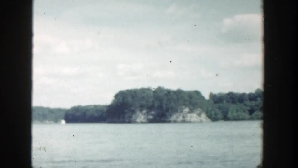 Faded 8mm frame of a Wisconsin lake in 1964, showing wooded shoreline, rocky outcrop, calm water, and a distant sailboat.