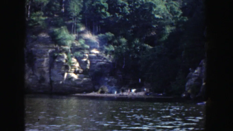 Wisconsin lake shoreline in 1964 with wooded rock cliffs, rippled water, and distant people gathered near the shore.