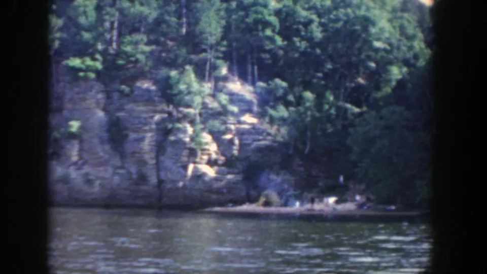A similar 1964 Wisconsin river view, with trees above a pale rock face and water filling the foreground.