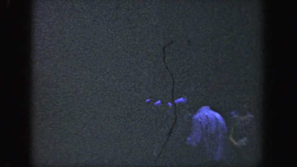 1964 Wisconsin 8mm frame showing a man low in the composition, partly hidden by darkness, grain, and tree shadows.