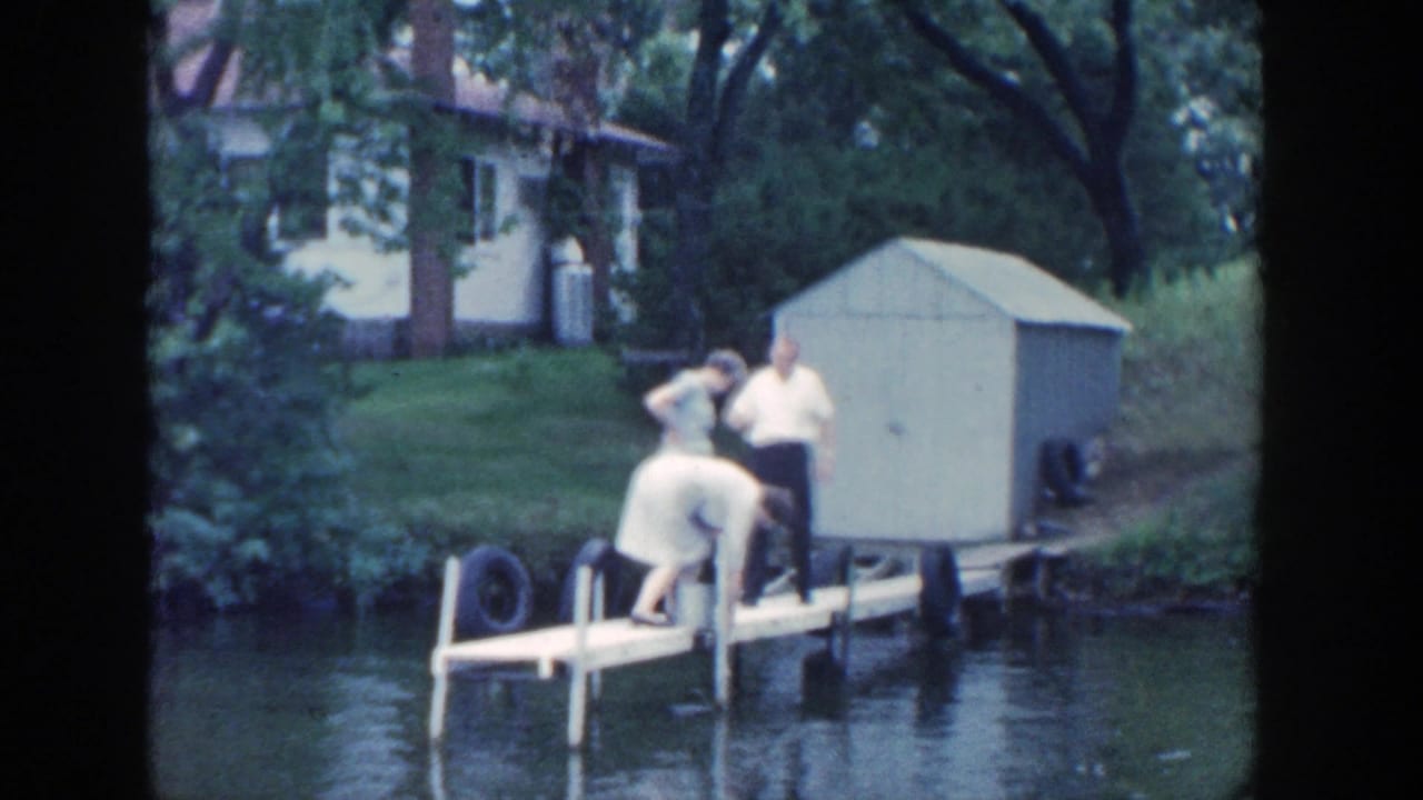 Adults stand on a wooden dock beside a calm river with a shed and house behind them.