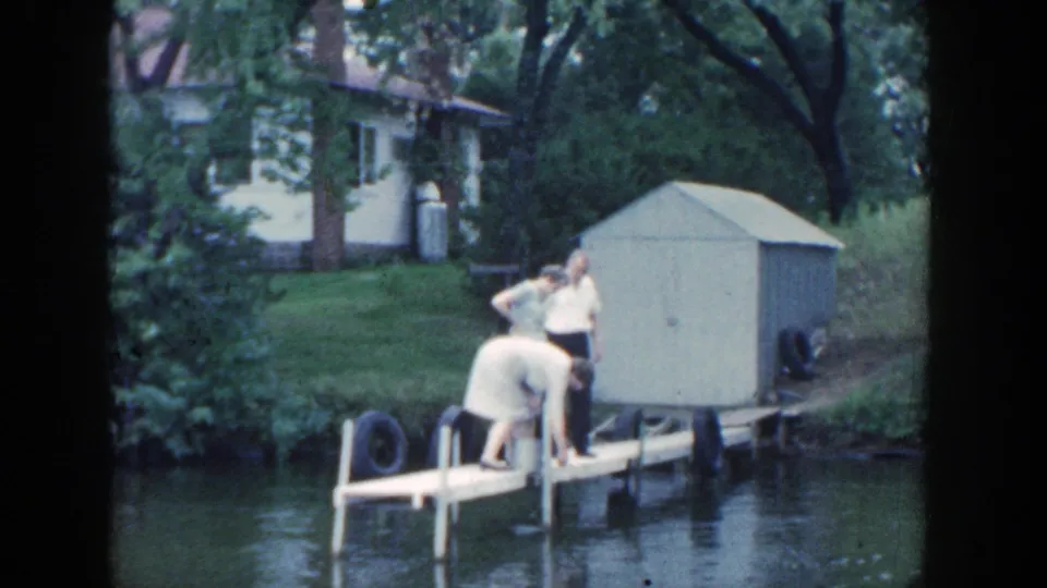 1964 Wisconsin frame of adults on a narrow dock over the river, with a pale shed, lawn, trees, and a white house visible behind them.