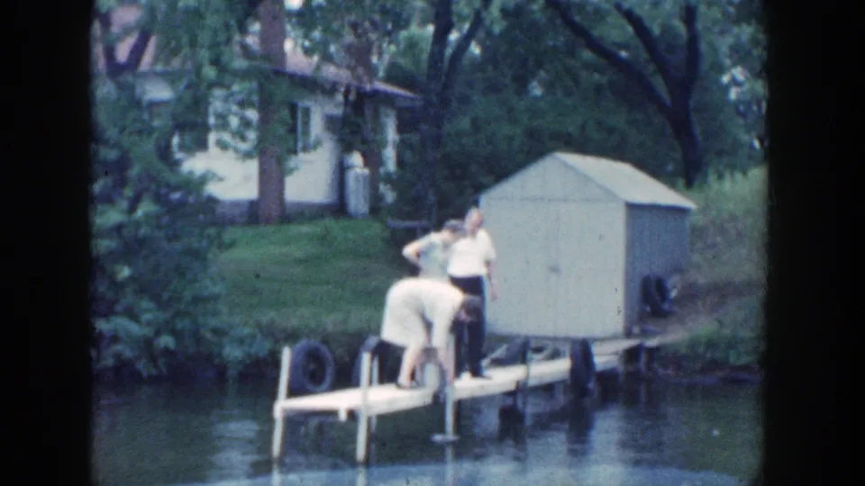 1964 Wisconsin home-movie view of family members on a dock by the river, framed with a small shed, white house, grass, and overhanging trees.