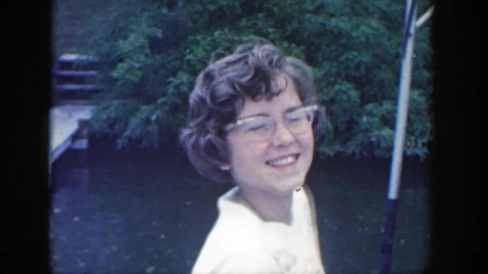 Close 1964 Wisconsin home-movie frame of a smiling young girl with curly hair and glasses beside the river, with leafy greenery behind her.