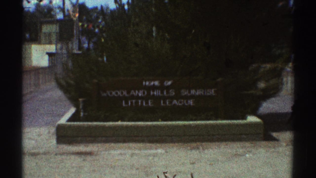Wide 1969 Woodland Hills frame showing the Little League sign partly distant, with bushes, walkway, and film-edge shadows visible.
