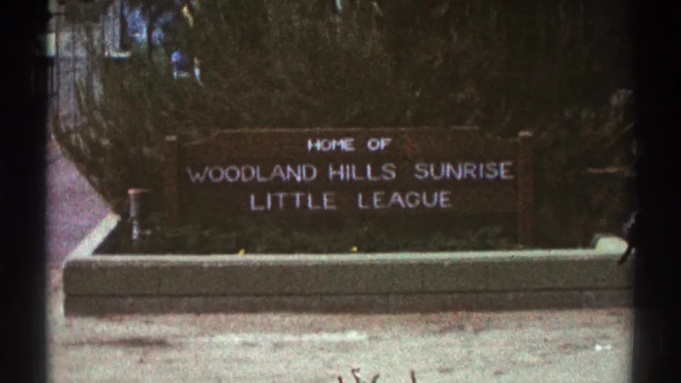 Mid-zoom 1969 Woodland Hills view of the brown Little League sign with white lettering, concrete base, and dense greenery behind it.