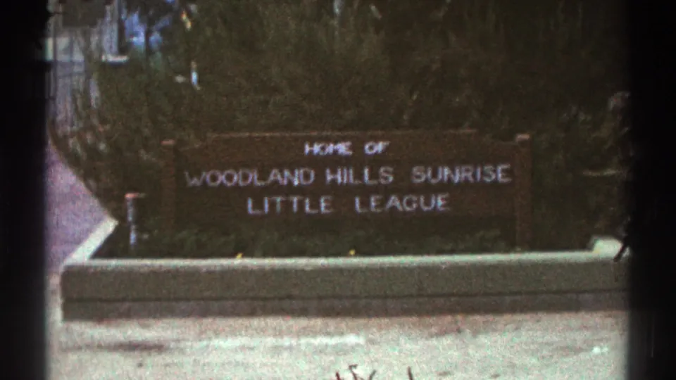 Closer 1969 home-movie frame of the Woodland Hills Sunrise Little League sign, with the concrete base and surrounding shrubs visible.