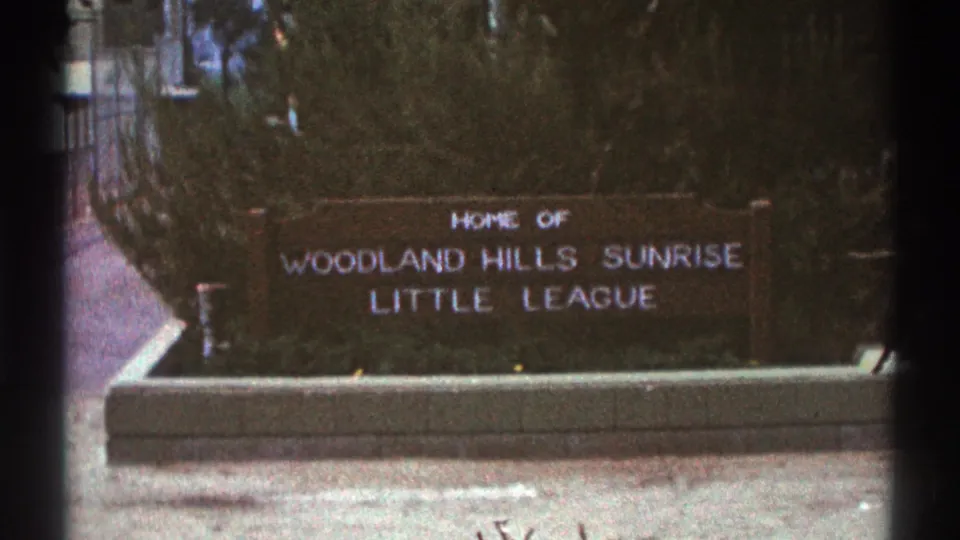 Final close 1969 Woodland Hills frame centers the Little League sign, with white lettering, bushes, and the low concrete base in view.