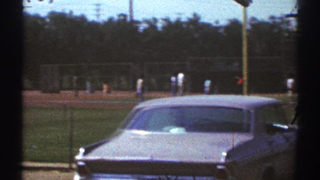 Parked sedan beside a chain-link fence with a baseball field in the background.