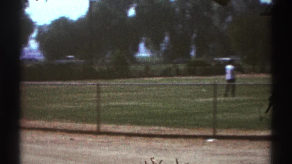 A distant figure in a white shirt stands on the public sports field, filmed from behind the fence in 1969 Woodland Hills.