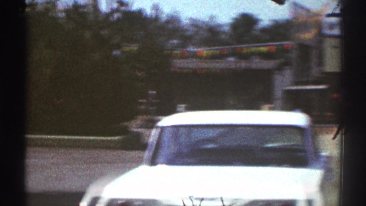 White mid-century car near a Woodland Hills sports field, with trees, bunting, and soft 8mm color fade visible.