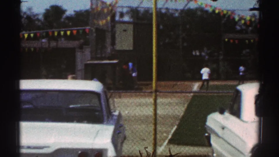 1969 Woodland Hills frame showing parked white cars, a narrow grass strip, distant people, and colorful pennants near the field.