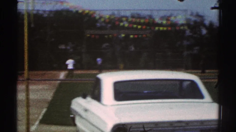 White car in the foreground at a 1969 Woodland Hills field, with two distant people, trees, and pennant bunting beyond.