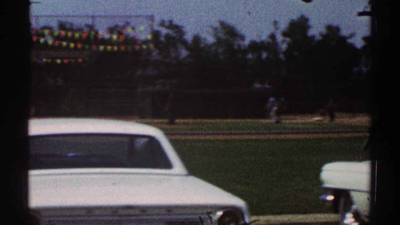 1969 Woodland Hills home-movie frame showing a white parked car, baseball field, pennant flags, trees, and distant players.