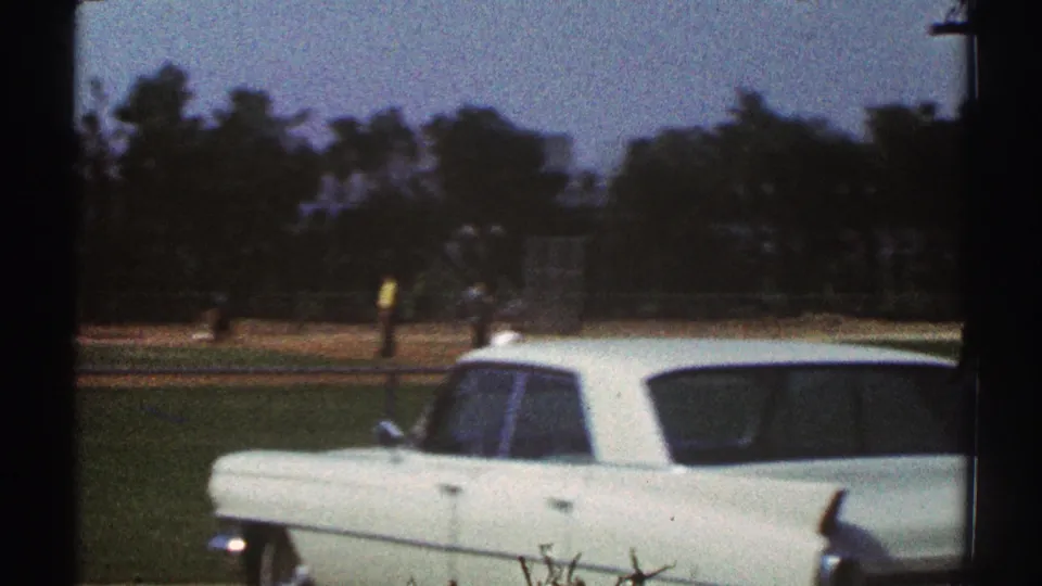 White station wagon parked near a Woodland Hills baseball field, with distant players and trees visible through soft 8mm grain.