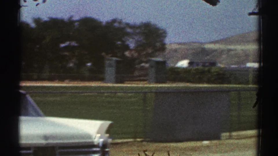 Home-movie frame from the baseball area showing part of a white car, field fencing, trees, and low hills beyond Woodland Hills.