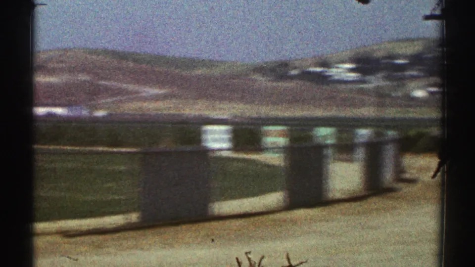 1969 Woodland Hills frame with low hills, open field edge, pavement, and parking-area posts seen from a roadside angle.
