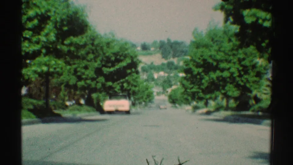 1969 Woodland Hills 8mm view down a sunny tree-lined street toward a parked pink car and distant hillside homes.