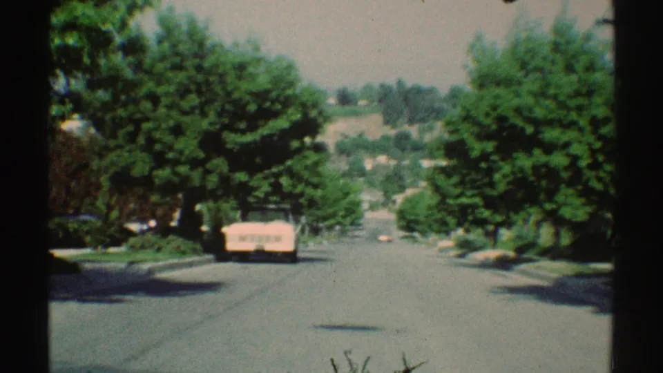 Approaching 1969 Woodland Hills street frame with a parked pink car, leafy trees, open roadway, and distant suburban hillside.
