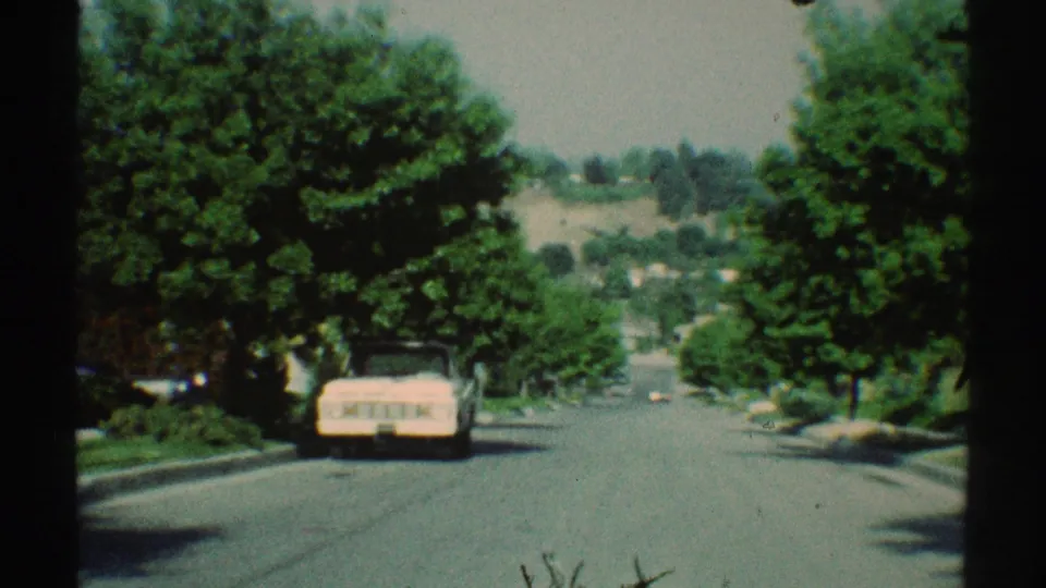 Late-clip 1969 Woodland Hills frame showing the parked pink car closer, green trees, sunlit pavement, and distant hillside suburb.
