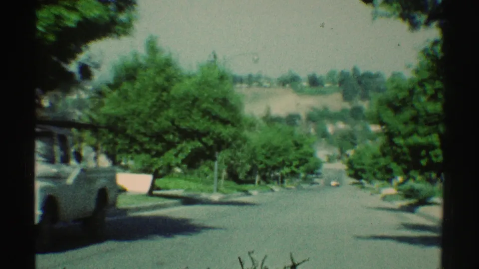 1969 Woodland Hills street view with a light vehicle at left, green residential landscaping, and distant hillside houses beyond the road.