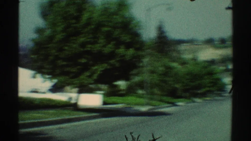 Blurred 1969 Woodland Hills 8mm frame showing a residential street edge, green trees, curb, lawn, and a pale house or wall at left.