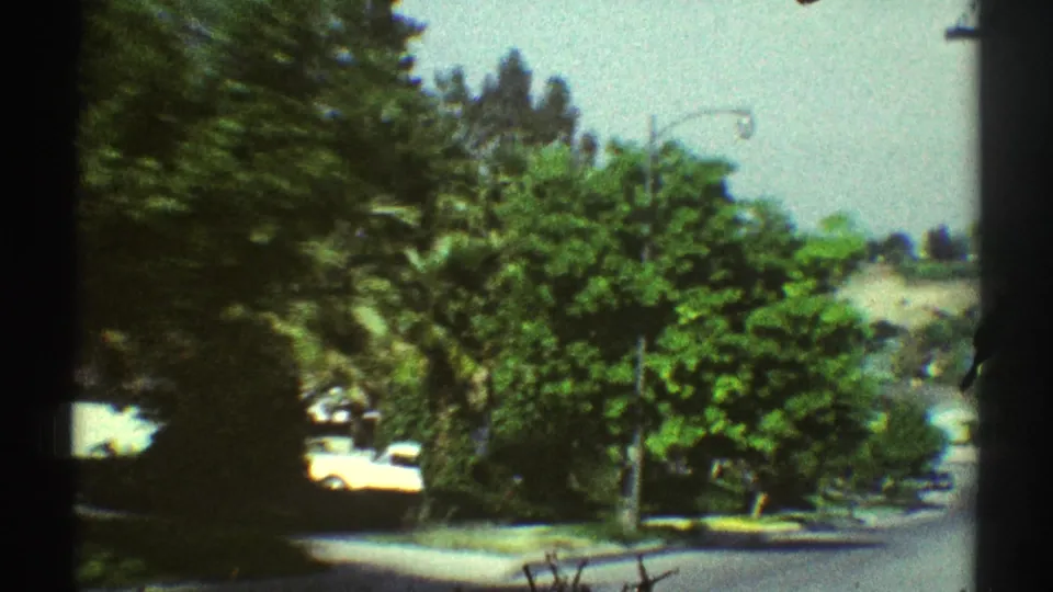 A downhill Woodland Hills street scene with mature trees, a streetlamp, sidewalk, parked cars, and hazy distant hills in vintage 8mm color.