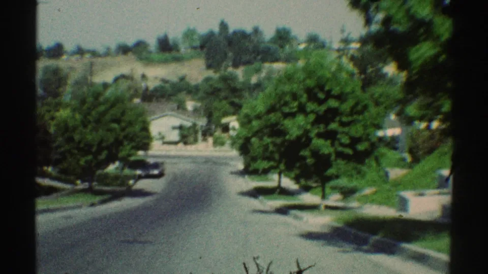 Approaching 1969 Woodland Hills residential street scene with a parked car, mature trees, lawns, and dry hillside beyond the houses.
