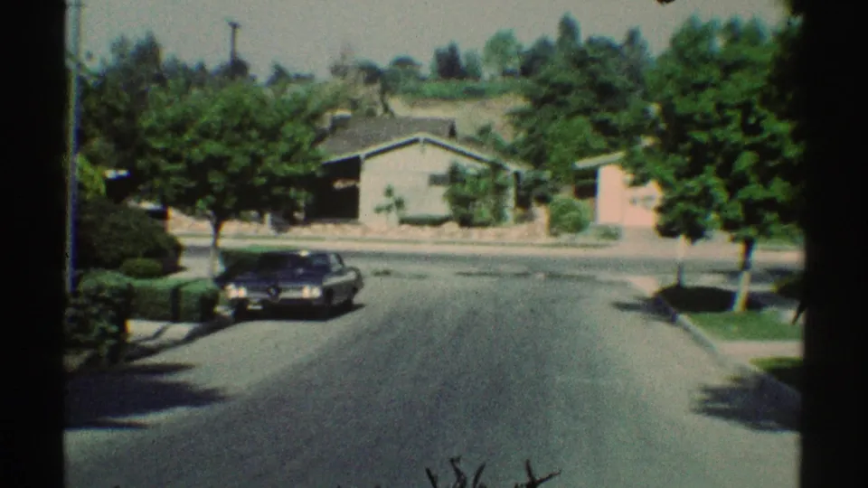 1969 Woodland Hills street scene from a moving vehicle, showing a dark sedan, shaded lawns, sidewalks, and single-story homes.