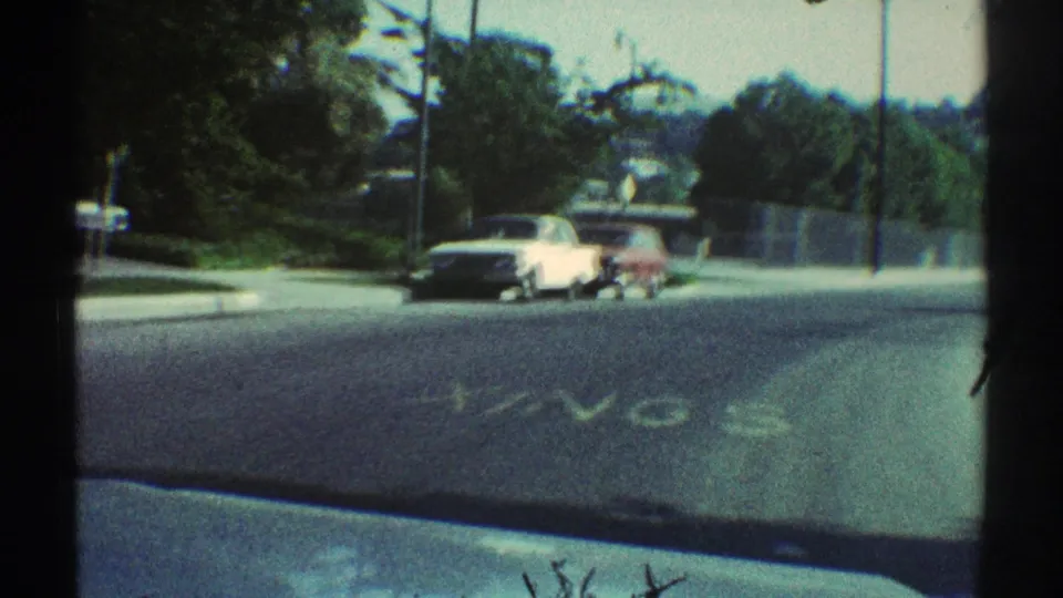 1969 Woodland Hills street scene with a white car and red vehicle near the curb, framed by dark window edges and a faded school road marking.
