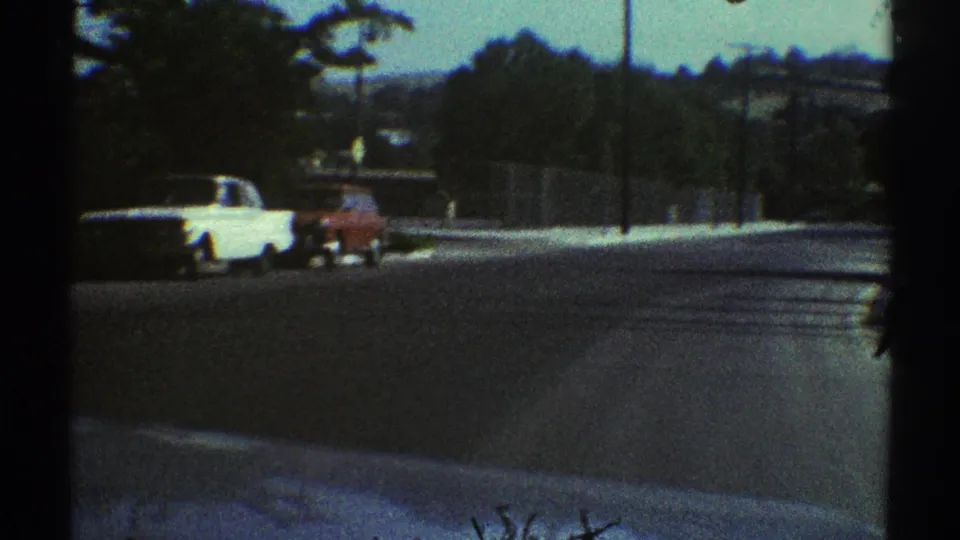 1969 Woodland Hills road view through a dark frame, showing a white car beside a red vehicle, fence line, trees, and shaded pavement.