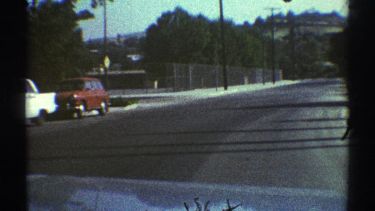 Suburban street seen through a windshield with a white car, red wagon, trees, poles, and distant hills.