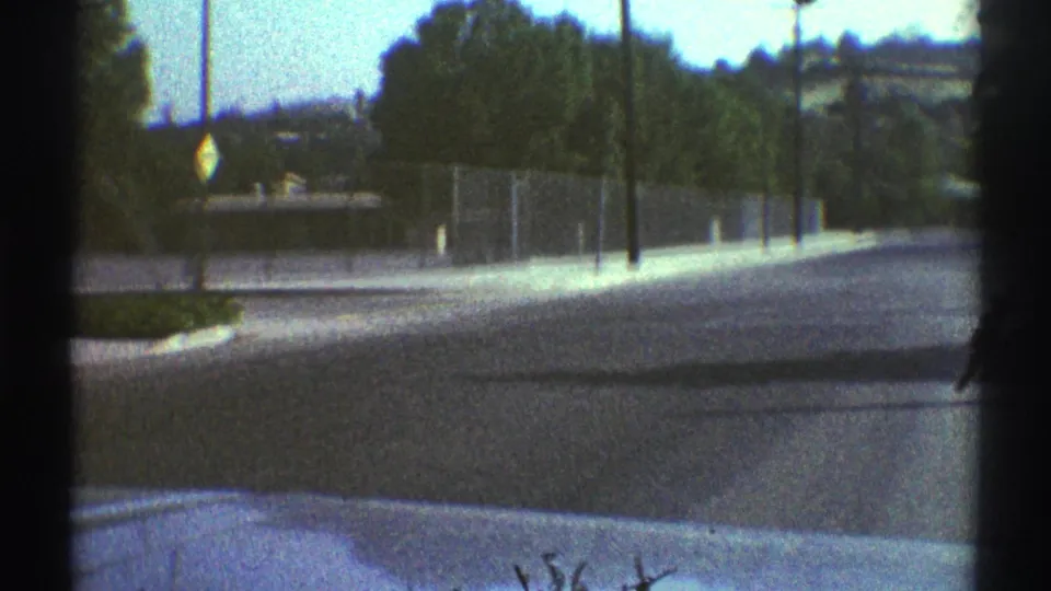 Woodland Hills roadway viewed from a moving car in 1969, with chain-link fencing, sidewalk, trees, street signs, and hazy hills.